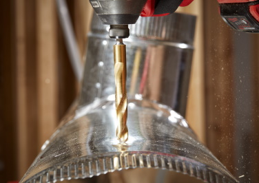 A Milwaukee power drill drives its drill bit through a galvanized heating duct.