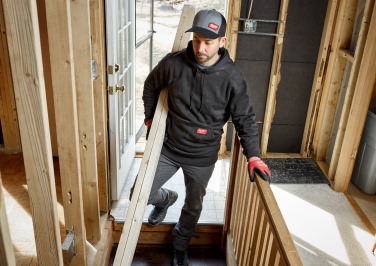 A workman carries an armful of 2x4 boards through a doorway while wearing a Milwaukee hoodie
