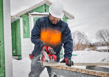 Workman in a heated Milwaukee jacket operates a circular saw in winter. His chest glows orange.